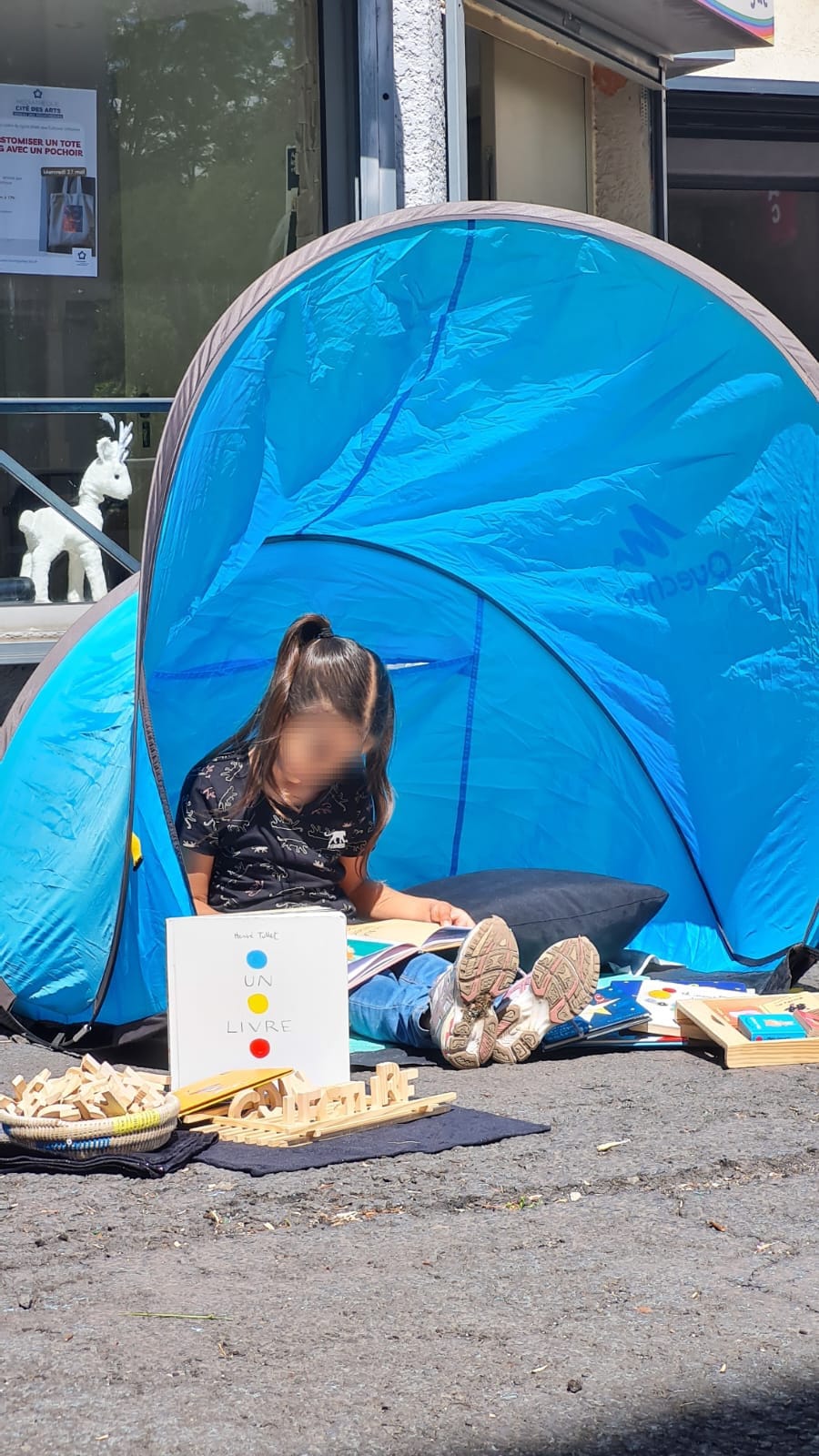 Petite tente installée sur un trottoir : un enfant y joue, livres et jeux posés au sol