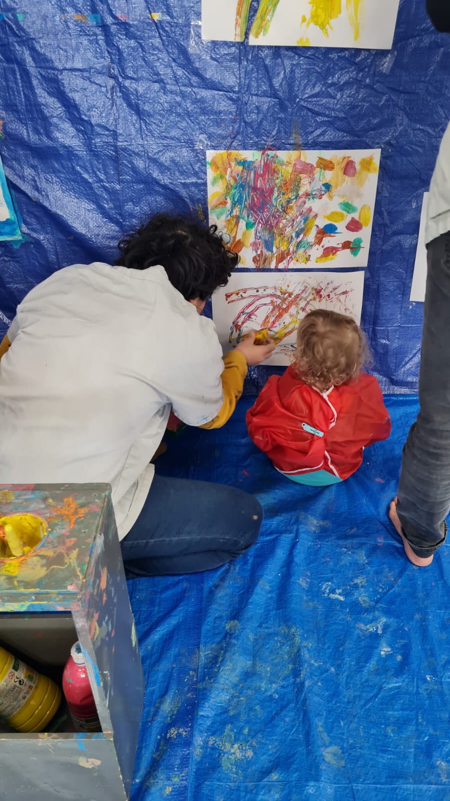 Une adulte accompagne un jeune enfant qui peint sur une grande feuille verticale